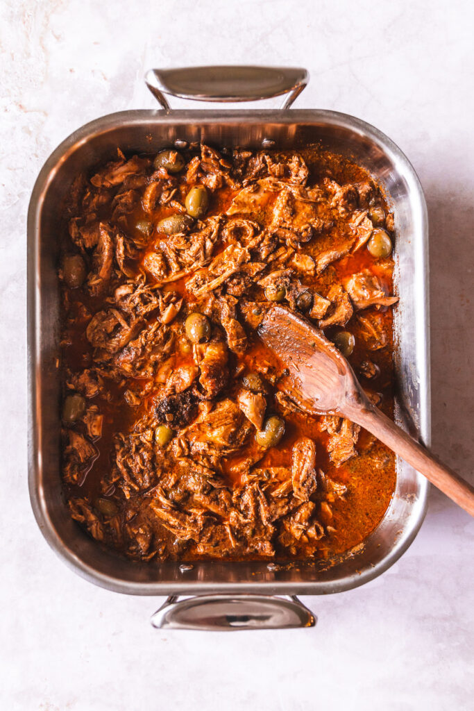 A roaster full of shredded turkey in a red sauce with green olives poking through