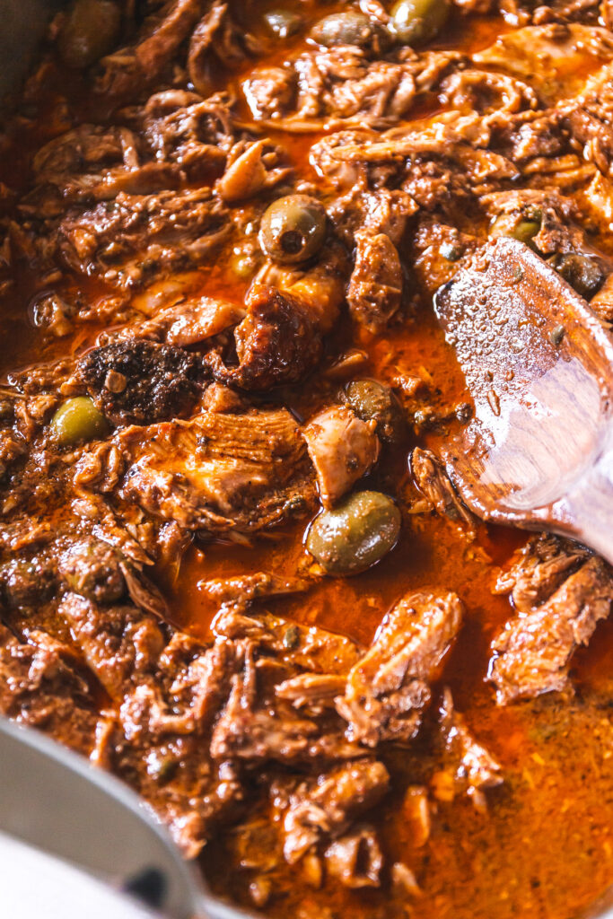 A roaster full of shredded turkey in a red sauce with green olives poking through