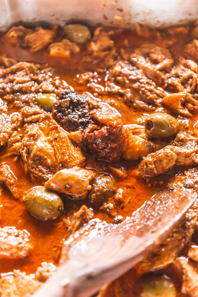 A roaster full of shredded turkey in a red sauce with green olives poking through