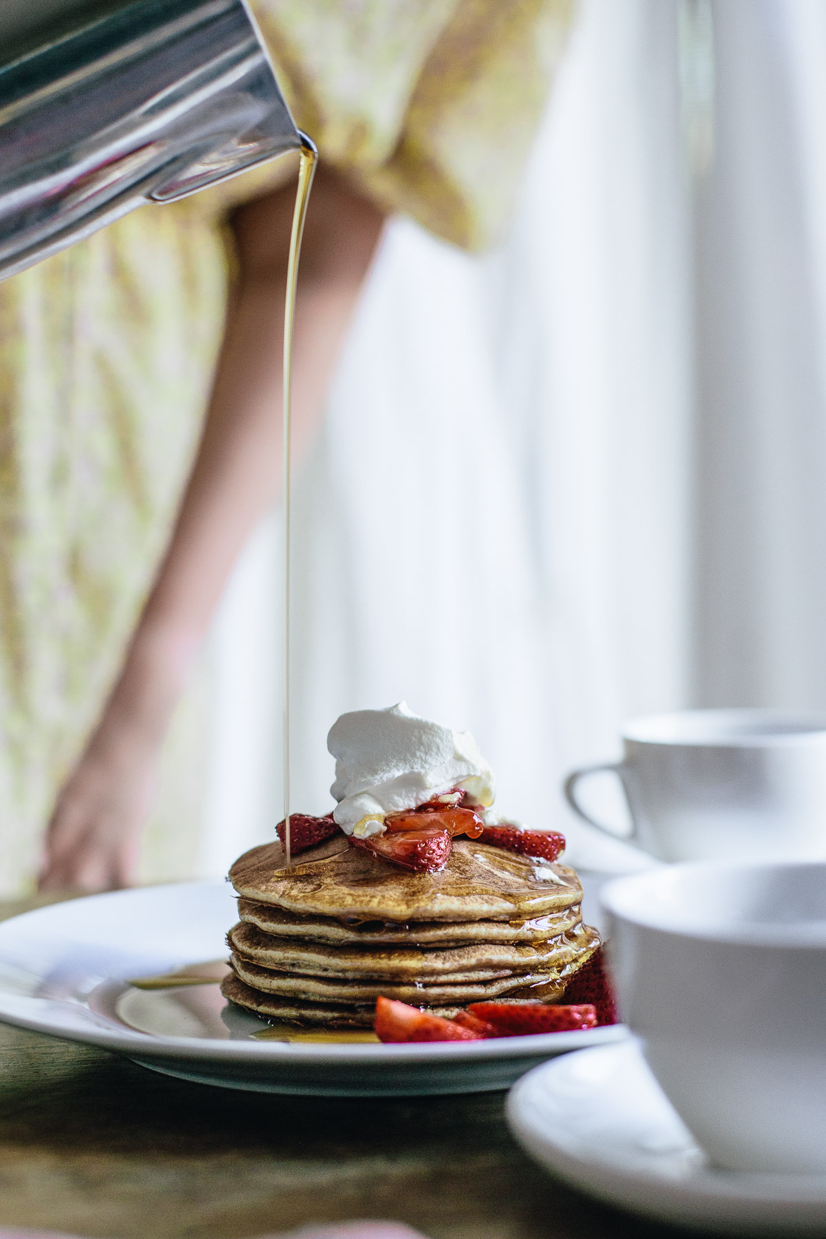 pancake stack with strawberries and whipped cream being poured with maple syrup