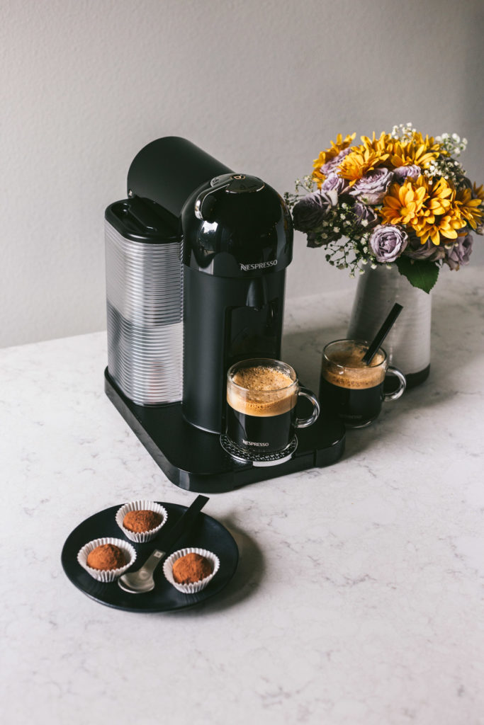 andgled image of a nespresso on a marbled surface with two cups of coffee, a bouquet of flowers and three truffles to the side