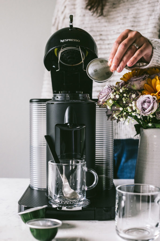 head on image of nespresso machine and a woman behind it placing in a new pod.