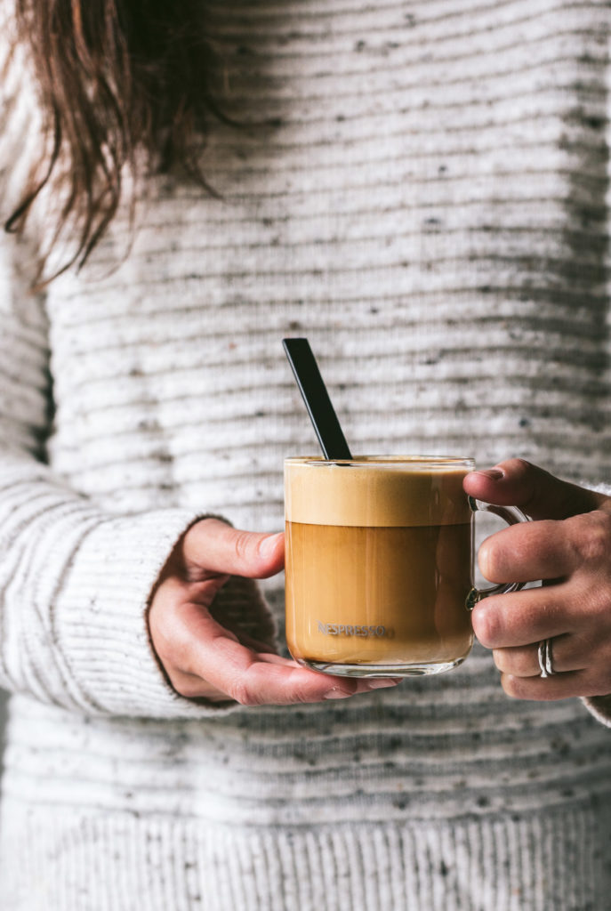 head on image of a woman holding a clear glass of milky coffee wearing a light grey sweater