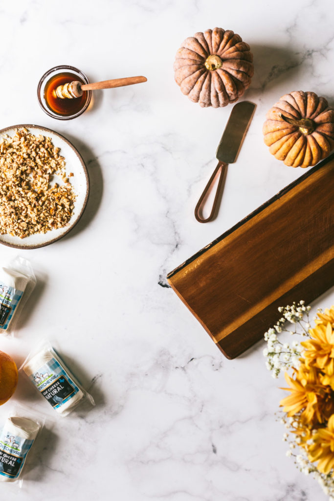 Overhead image of ingredients on a marble countertop: thin, dark cheese board. Montchevre goat cheese, toasted walnuts, honey, and decorative small pumpkins