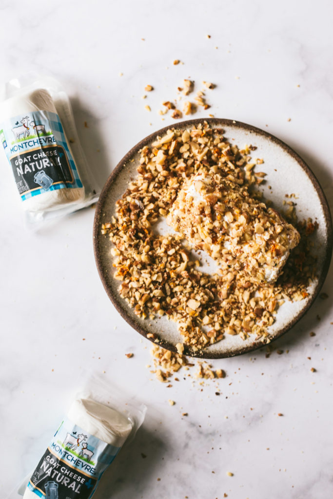 Overhead image on a marble surface. Toasted walnuts on a plate and opened goat cheese bing coated in toasted walnuts