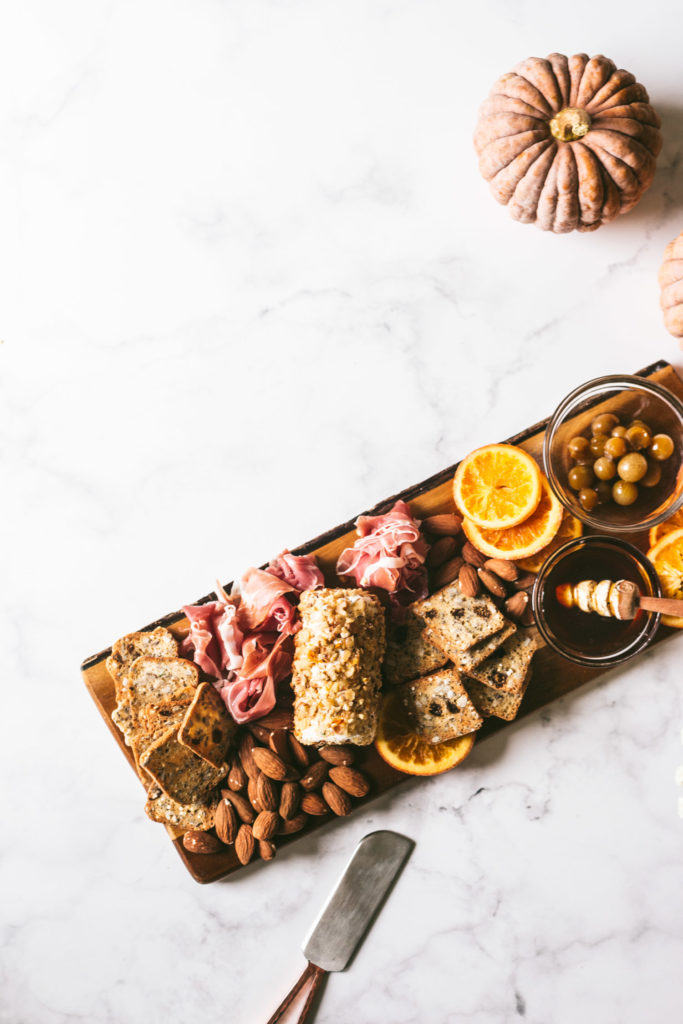 Overhead image of plated charcuterie board on marble surface. From left to right, herbed crackers, almonds, procuitto, Toasted walnut goatcheese, herbed crackers, candied oranges, honey, olives