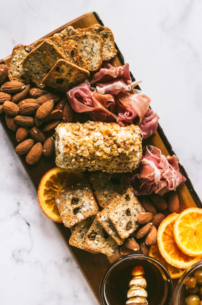 Overhead image of plated charcuterie board on marble surface. From top to bottom, herbed crackers, almonds, procuitto, Toasted walnut goatcheese, herbed crackers, candied oranges, honey, olives