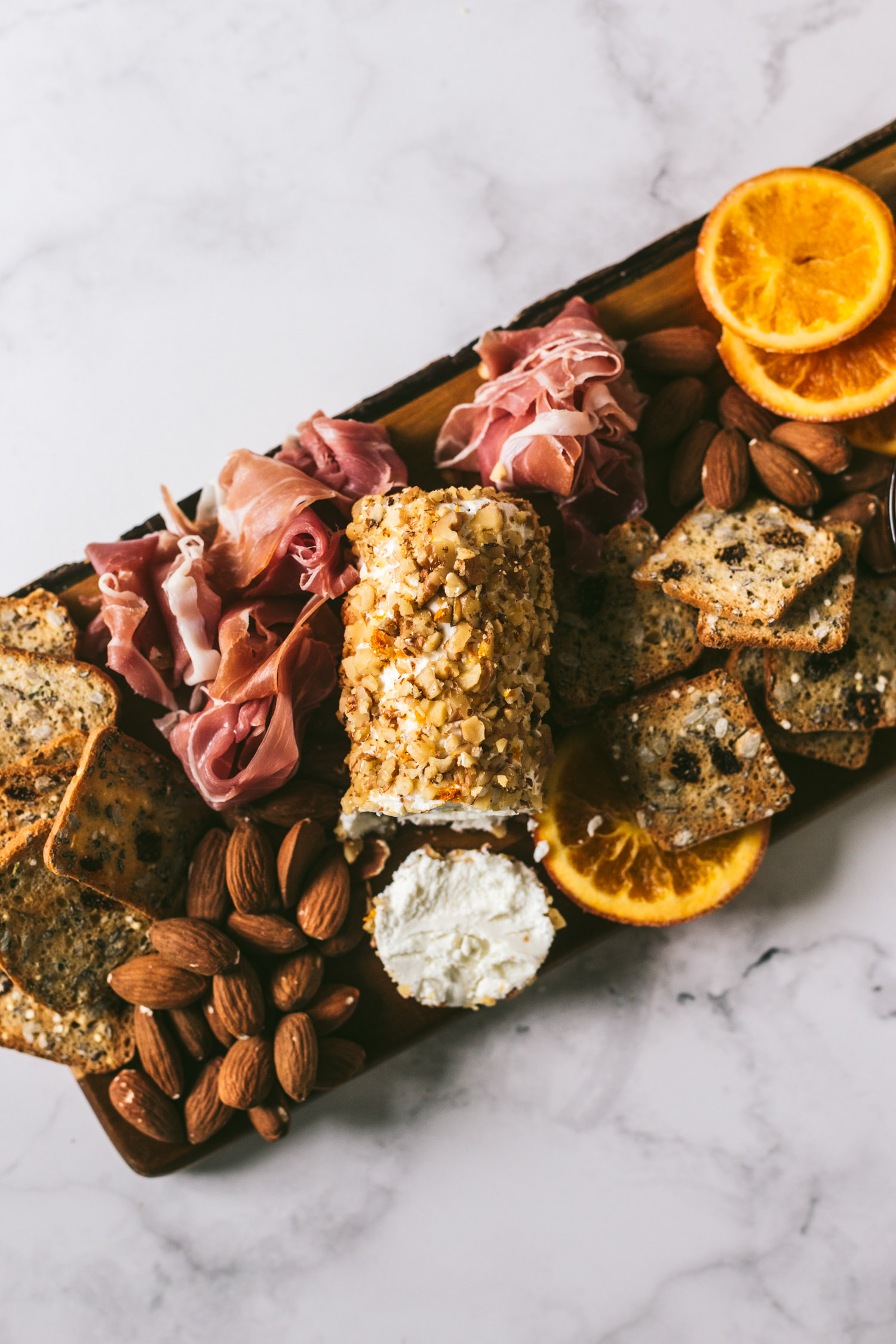 Overhead image of plated charcuterie board on marble surface. From left to right, herbed crackers, almonds, procuitto, Toasted walnut goatcheese, herbed crackers, candied oranges, honey, olives