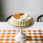 Orange spice cake on top of a tall white cake stand sitting on a marbled table with a mustard checkered runner