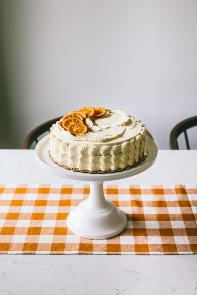 Orange spice cake on top of a tall white cake stand sitting on a marbled table with a mustard checkered runner