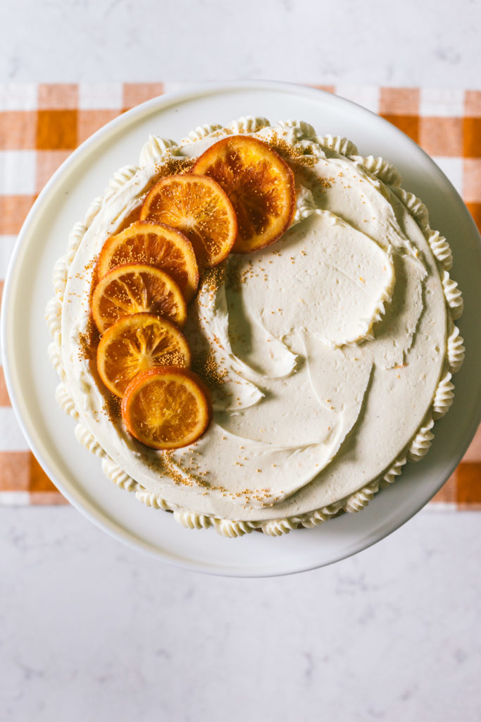 Top down image of Orange spice cake on top of a tall white cake stand sitting on a marbled table with a mustard checkered runner. Cake is topped with candied oranges off to the side, speckled with gold sprinkles and the frosting is swirled around.