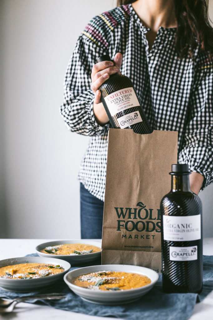 Head on image of a girl lifting a bottle of carapelli olive oil out of a whole foods paper bag. Bowls of Butternut Squash Soup to her right.