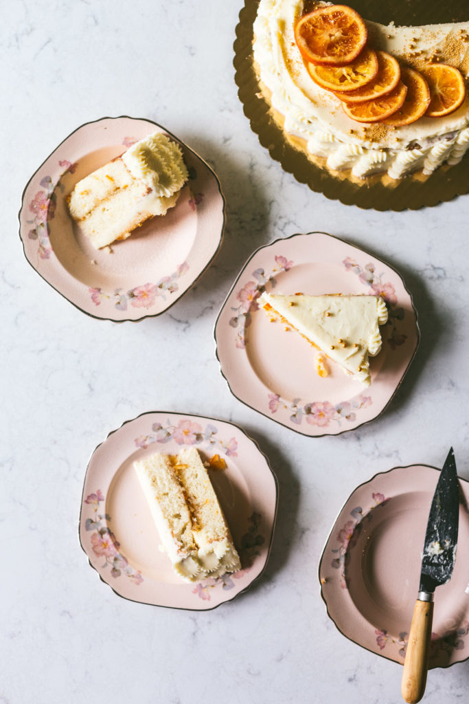 4 vintage pink plates hold the Orange spice cake sliced onto three plates. One plate on the bottom left has a wooden handled knife. The top right corner has the rest of the cake just peeking through the image.