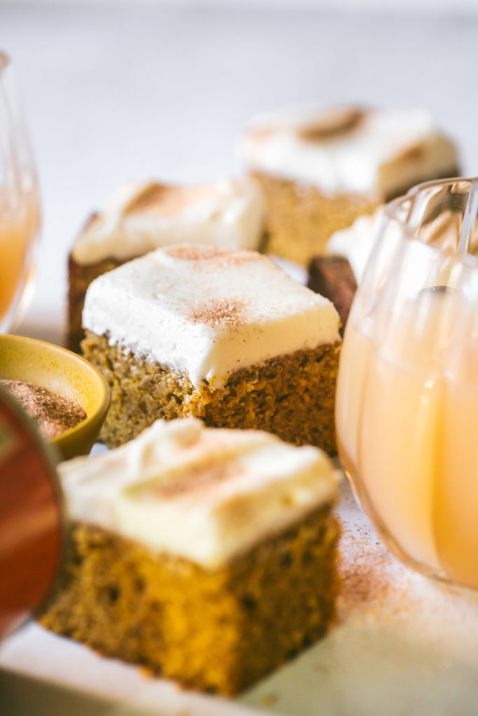 Pumpkin Bars with Browned Butter Cream Cheese Frosting sprinkled with cinnamon and sugar cut into squares next to apple cider