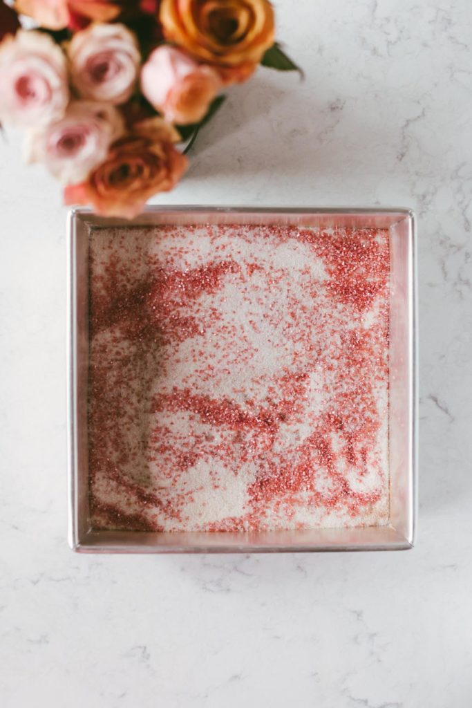 Overhead image of pink sugar in a square shaped metal pan with flowers off to the left.