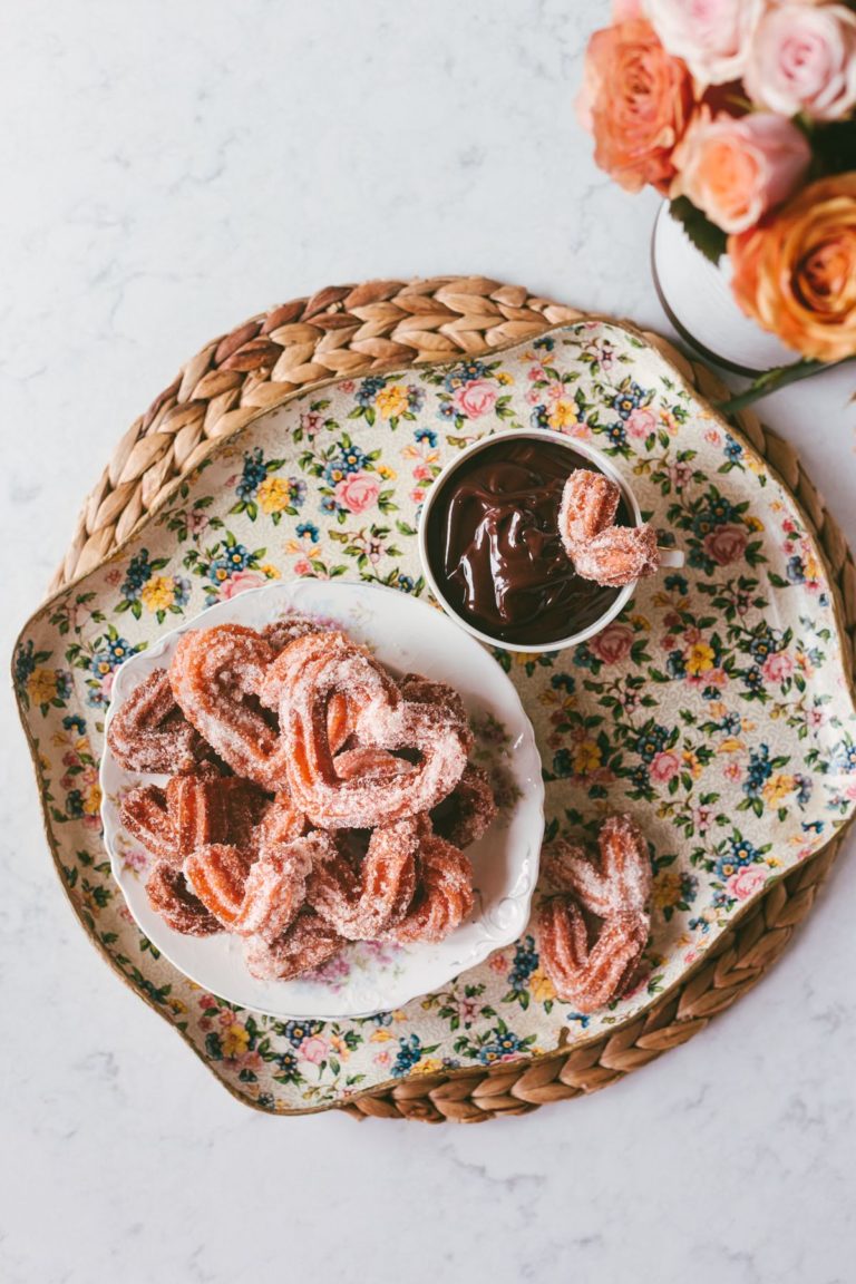 Over head shot of Pink Heart-Shaped Churros in a bowl, next to a bowl of ganache and a vase of pink roses.