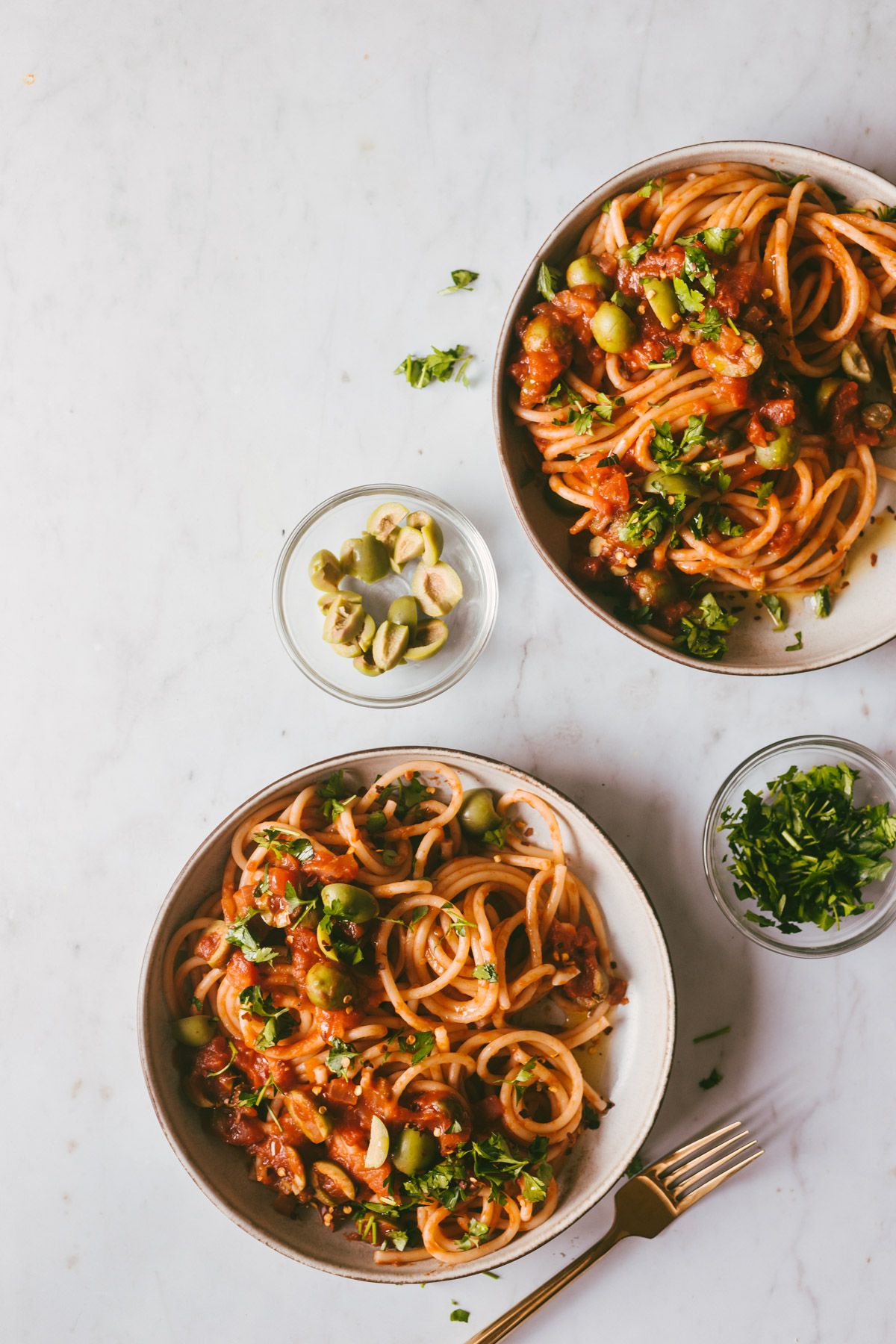 Two bowls of finished Pasta Puttanesca with extra olives and parsley on the side.