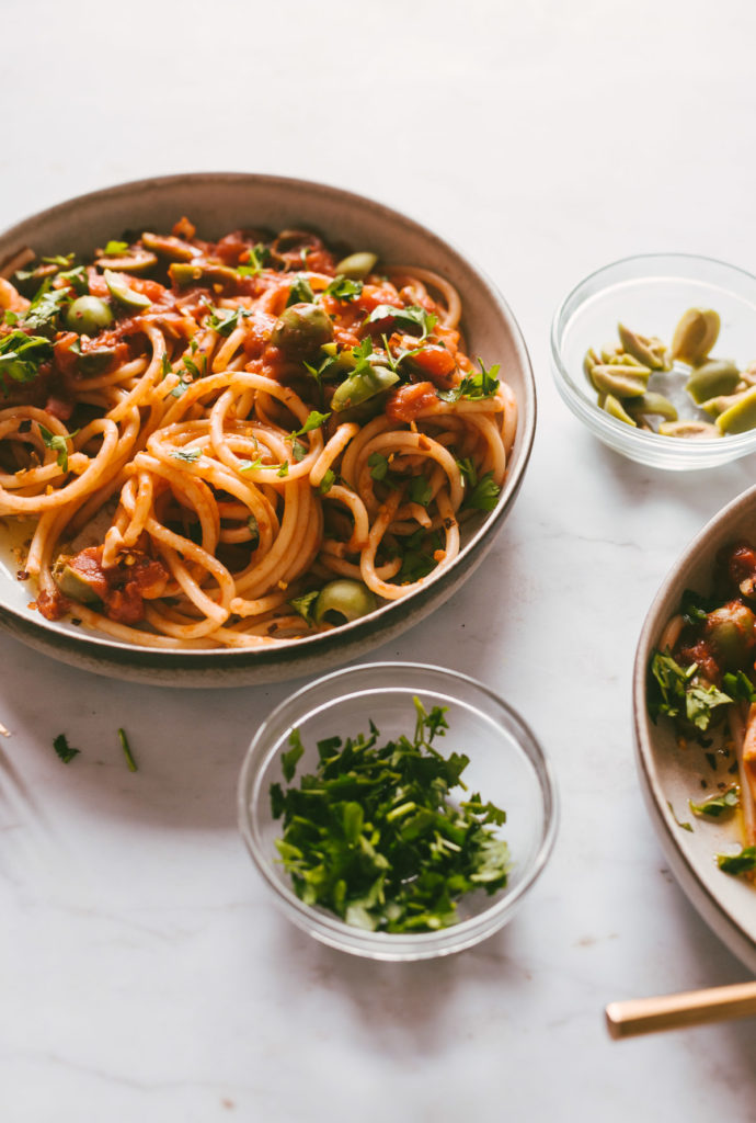 Two bowls of finished Pasta Puttanesca with extra olives and parsley on the side.