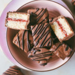 The square petit four in a pink bowl and off to the sides with one petit four being cut open revealing the strawberry center.