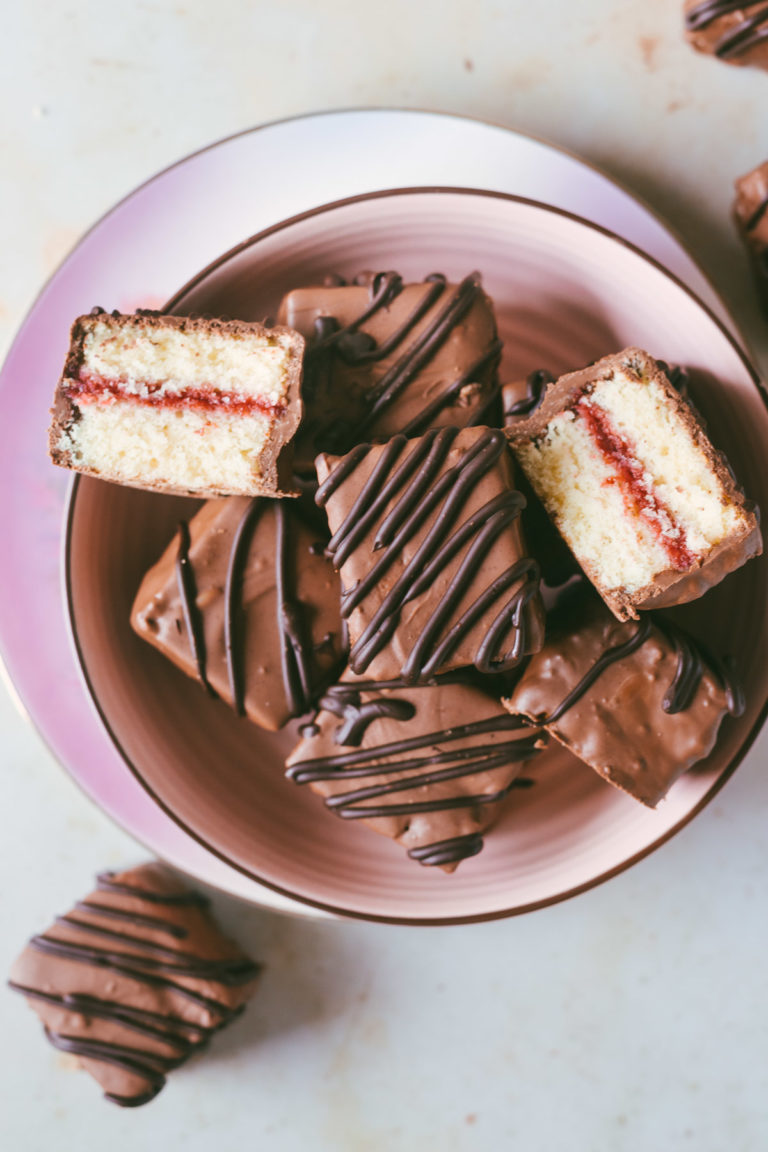 The square petit four in a pink bowl and off to the sides with one petit four being cut open revealing the strawberry center.