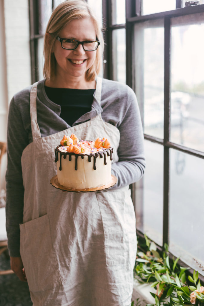 A woman dressed in black holding her decorated cake with chocolate drip