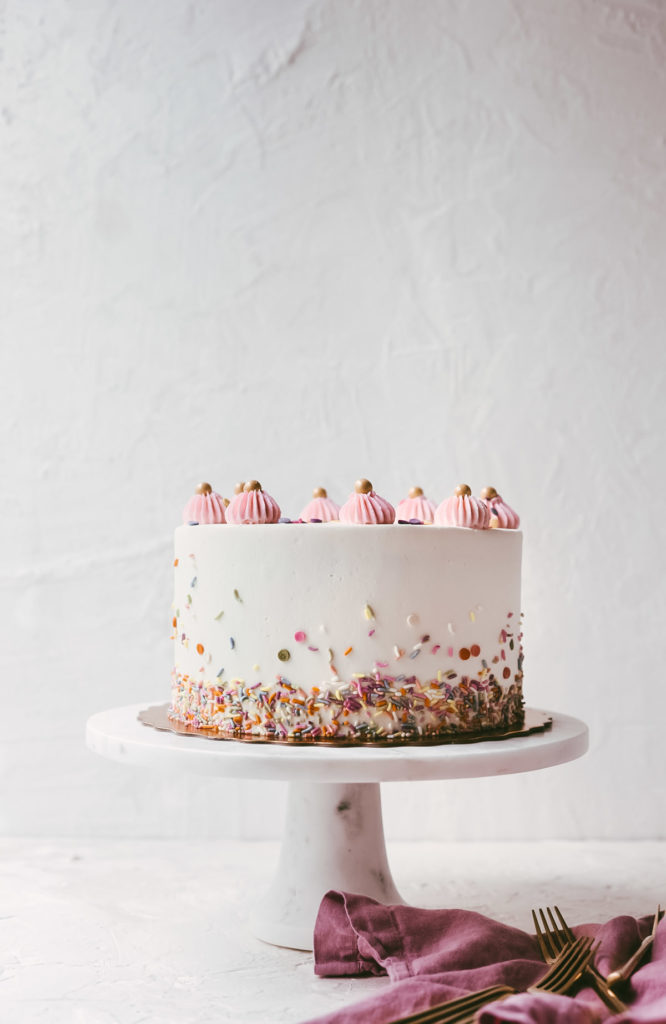 Head on image of a white cake with sprinkles up the sides and pink buttercream dollops on the top