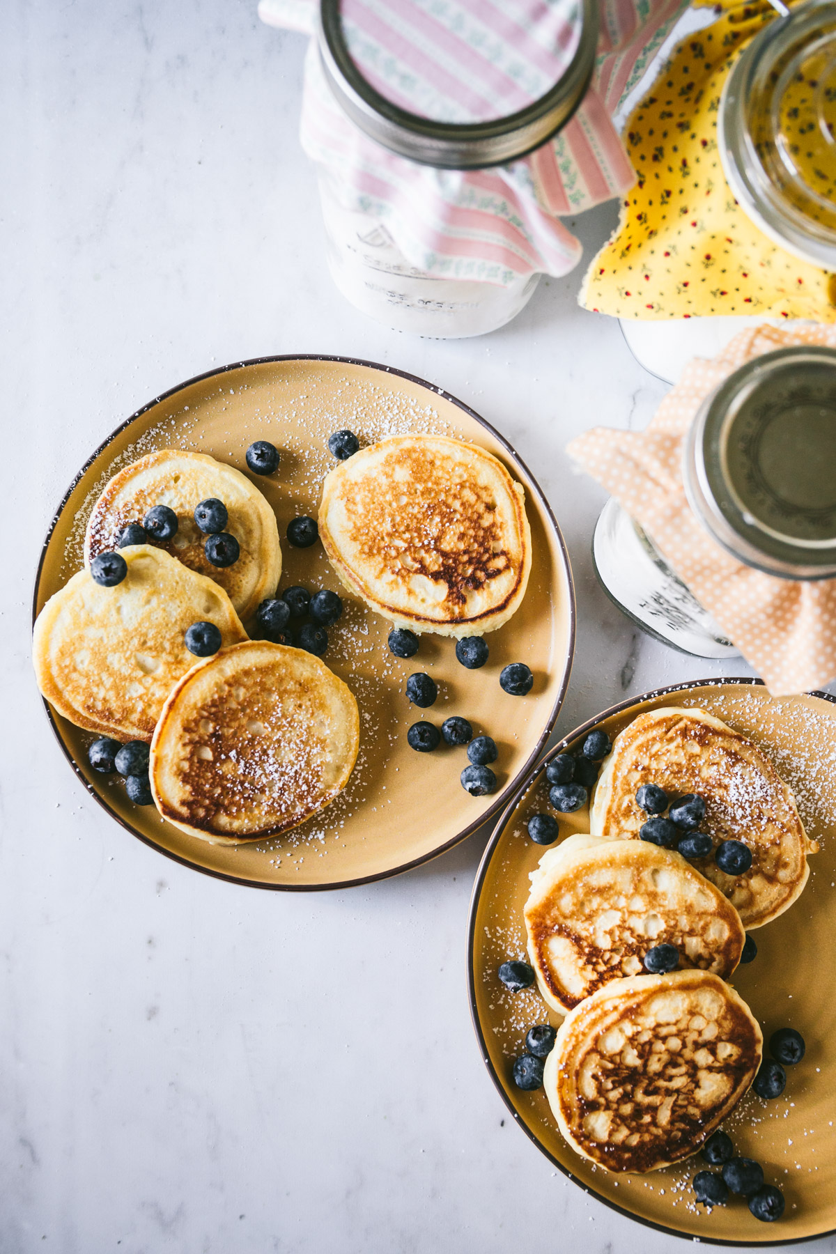 Overhead image of two mustard colored plates with small pancakes on top sprinkled with blueberries. The buttermilk pancake mix is on the upper right hand side.