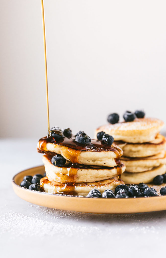 head on image of two stacks of pancakes, with bluberries everywhere and maple syrup being drizzled over the top