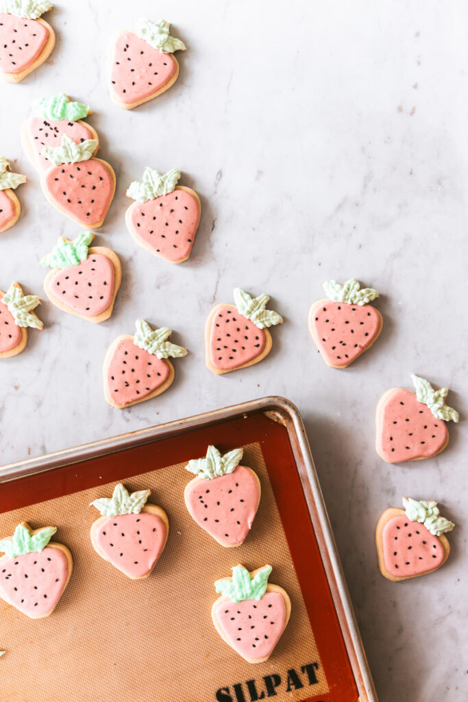 Overhead picture of strawberry shaped cookies with pink icing, black sesame seed decorations and green frosting leaves