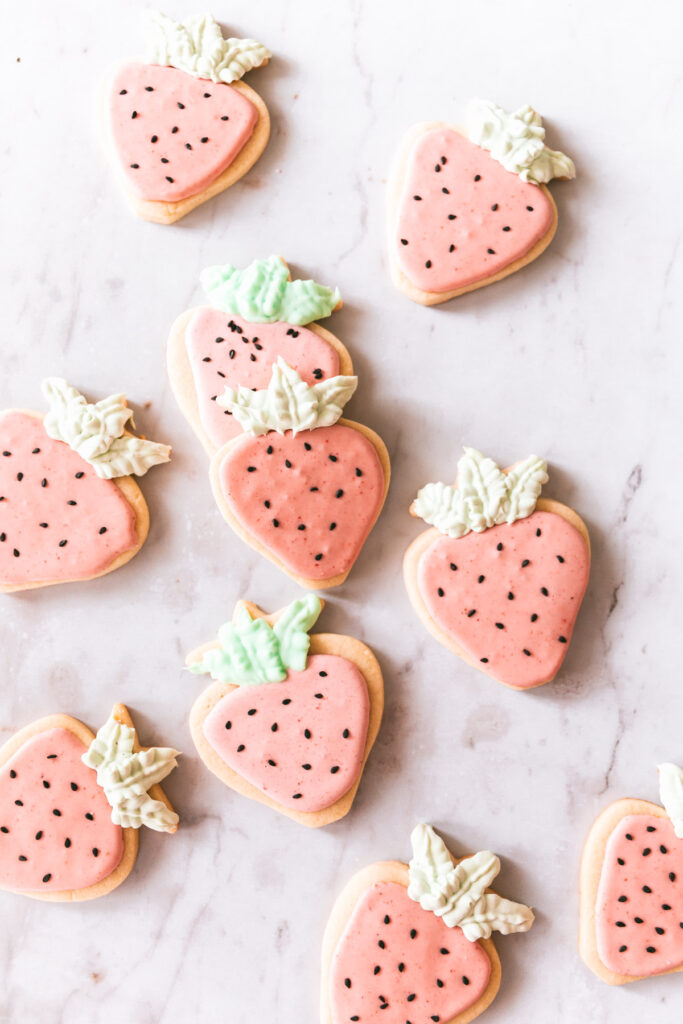 Overhead picture of strawberry shaped cookies with pink icing, black sesame seed decorations and green frosting leaves