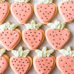 Overhead picture of strawberry shaped cookies with pink icing, black sesame seed decorations and green frosting leaves