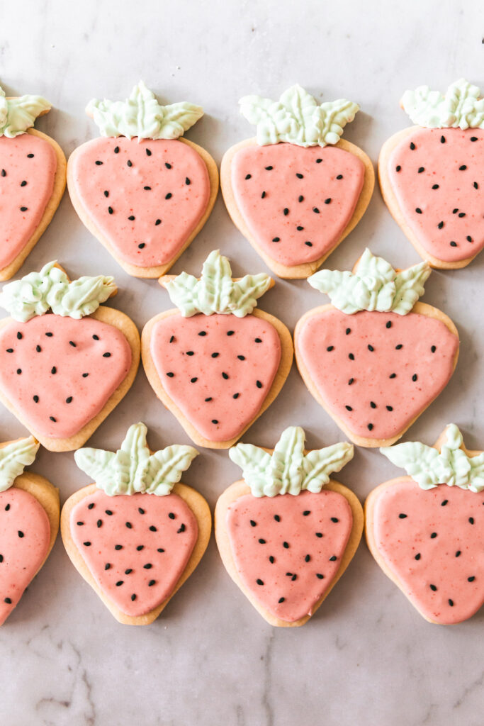 Overhead picture of strawberry shaped cookies with pink icing, black sesame seed decorations and green frosting leaves