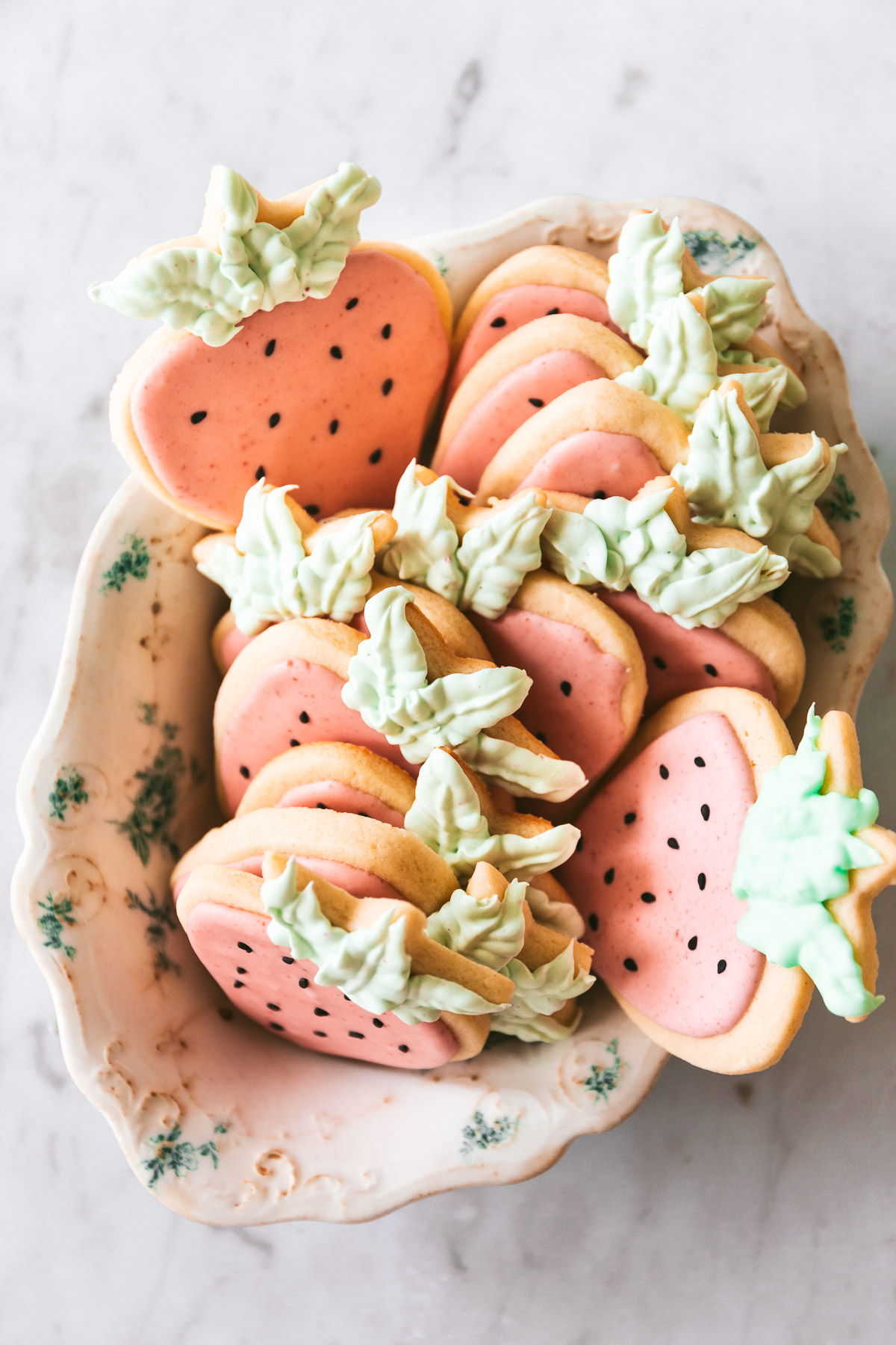 Overhead picture of strawberry shaped cookies with pink icing, black sesame seed decorations and green frosting leaves in a vintage bowl