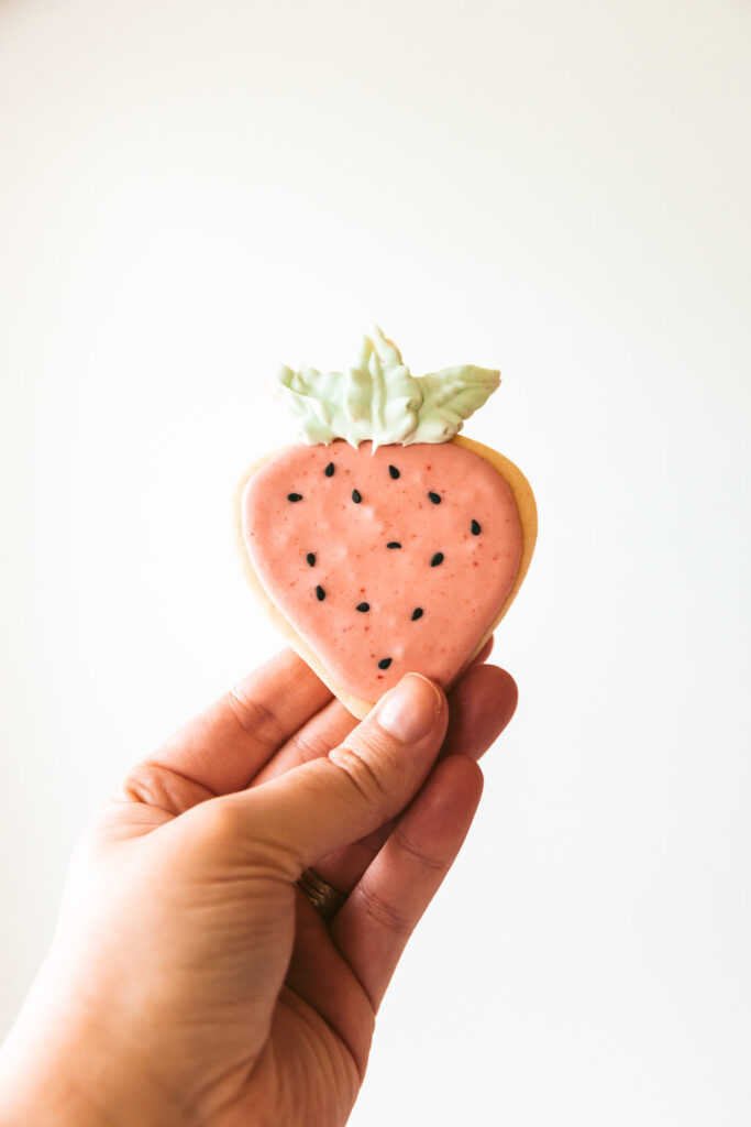 hand holding a strawberry shaped cookie with pink icing, black sesame seed decorations and green frosting leaves