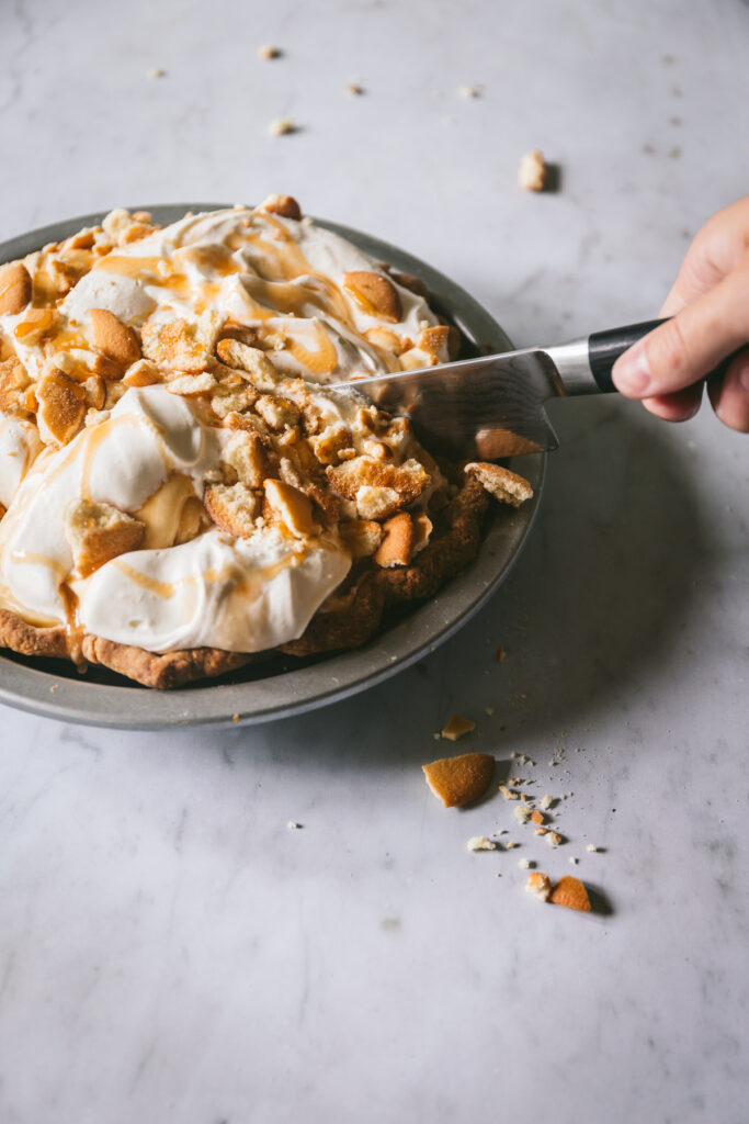 Image of full pie with soft whipped cream, caramel drizzle and crushed nilla wafers being cut with a knife