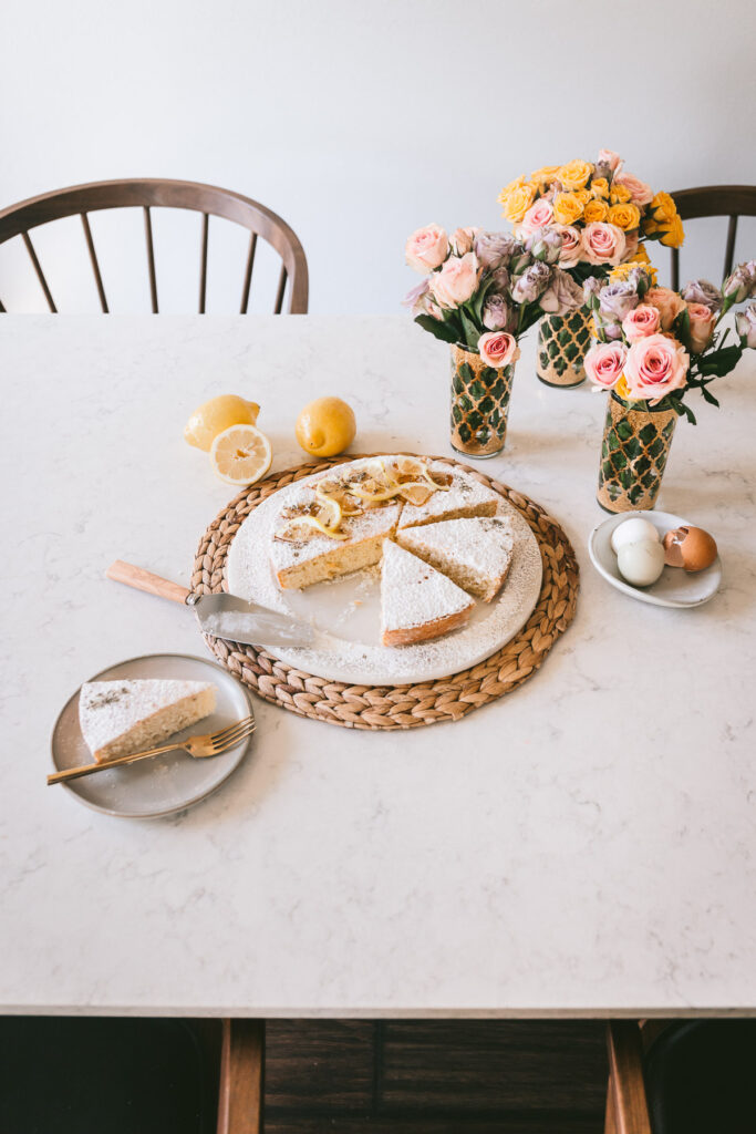 Head on image of the Head on angle of cake with a slice slightly moved away from the cake and cake slices on cake plates on a marble table with walnut chairs.