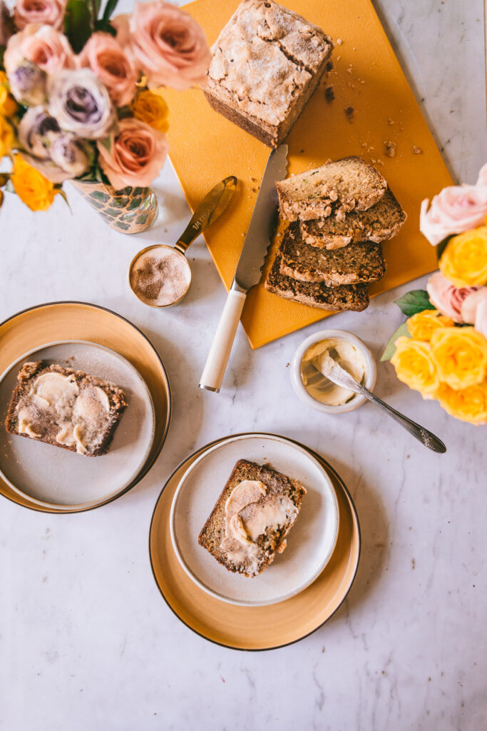 overhead image of zucchini bread slices on plates, buttered and sugared with florals on the table, knives and other props